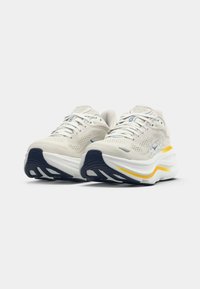 Scarpe sportive bianche con tomaia in rete traspirante, suola in gomma blu navy e dettagli gialli sulla soletta. Progettate per comfort e supporto.