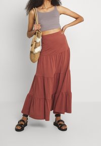 Jupe maxi évasée marron en tissu doux, associée à un top court gris. Un sac en osier et des sandales noires complètent la tenue.