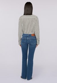 Mujer con cabello oscuro y lacio, vistiendo una camiseta de rayas de manga larga metida dentro de unos jeans azules de cintura alta, de pie de espaldas sobre un fondo liso.