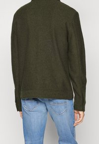 Olivgrön ulljacka med krage och långa ärmar, med en något texturerad yta och skräddarsydd nederkant, i kombination med ljusblå jeans.