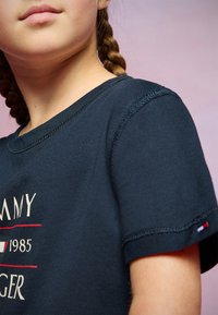 Námořnické bavlněné tričko s kulatým výstřihem, krátkými rukávy a grafickým potiskem s červenými a bílými akcenty. Text zní "Tommy 1985."