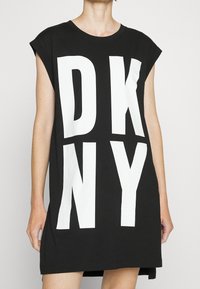 Svart bomulls t-shirt klänning med vit "DKNY" tryck. Avslappnad passform, korta ärmar, rund halsringning och fållen är något längre bak.