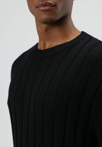 Pull noir côtelé avec un col rond, présentant des motifs verticaux texturés et un tissu en maille douce. Gros plan sur le haut du torse.