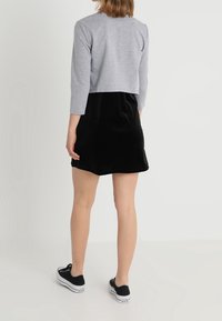 Femme debout, portant un pull gris court, une jupe noire et des baskets noires à tige basse, tournant le dos contre un fond simple.