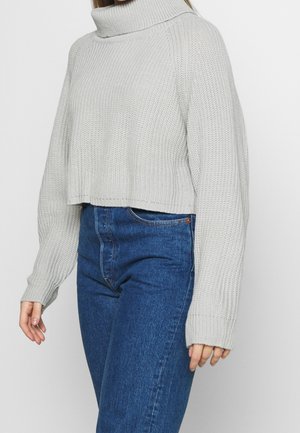 Harmaa lyhyt villapaita, jossa on korkea korkeakaula, ribbitetty pinta ja leveät hihat, yhdistettynä korkeavyötäröisiin sinisiin farkkuihin.