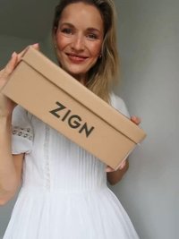 Braune Pappschuhschachtel mit geprägt "ZIGN" Logo, gehalten von einer Person, die ein weißes, plissiertes Kleid mit Knopfdetails trägt.