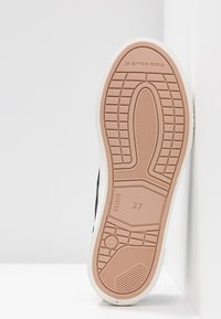 Semelle de sneaker noire en matériau en caoutchouc, surface texturée et accents beiges. Présente un logo G-Star RAW et une indication de taille 37.