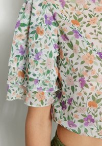 Top corto floreale con maniche a strati e volant, realizzato in tessuto leggero e trasparente su sfondo bianco, decorato con motivi arancioni, viola e verdi.