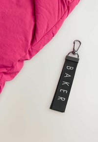Rozā siltināta auduma ar uzsūcošu tekstūru un pūkaina dizaina, kopā ar melna auduma tagu, uz kura baltajās burtos ir rakstīts "BAKER", un sudraba klips.