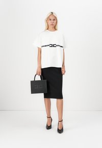 White short-sleeve top with black logo belt, paired with a knee-length black skirt. Black studded handbag with handles. Black pointed heels.