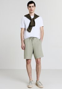 Jeune homme debout, portant un t-shirt blanc, un short vert clair, des chaussures beiges et une chemise olive nouée autour des épaules.