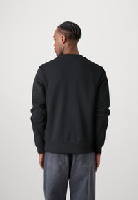 Felpa nera con scollo rotondo, maniche lunghe e polsini a coste. Il tessuto appare liscio e spesso, abbinato a pantaloni in denim grigi.