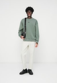 Green sweatshirt with round neck, black logo, paired with white pants and black sneakers. Accessories include a small black crossbody bag.