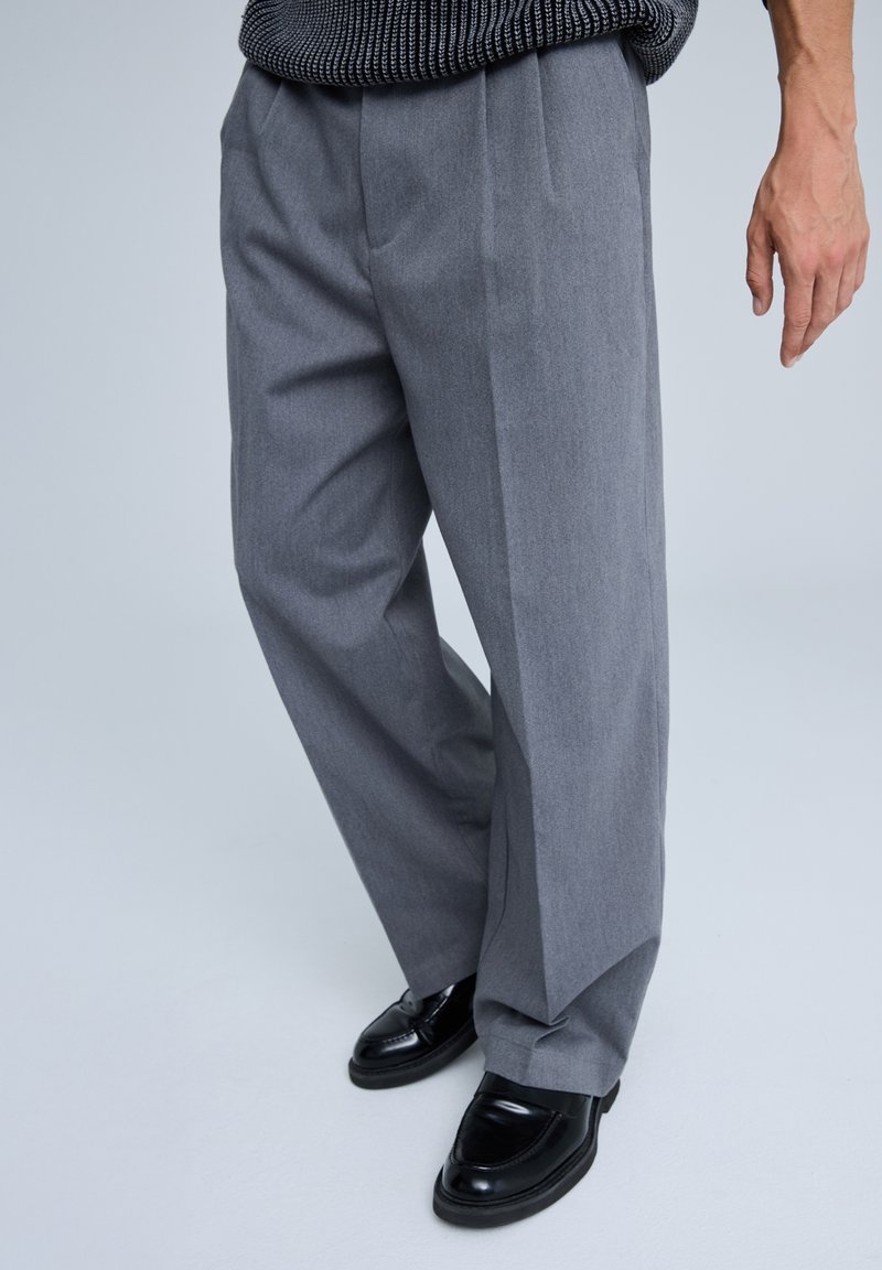 Pantaloni grigi su misura con un design a gamba dritta, caratterizzati da pieghe, una texture liscia e indossati con scarpe eleganti nere lucide.