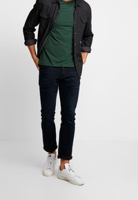 Herreoutfit med en mørk knap-op skjorte over en grøn t-shirt, parret med mørke jeans og hvide sneakers. Afslappet stil med opkrøllede ærmer.