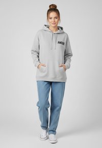 Felpa leggera grigia con cappuccio e stampa "AMOUR", tasche frontali e polsini a costine, abbinata a jeans blu e sneakers bianche.