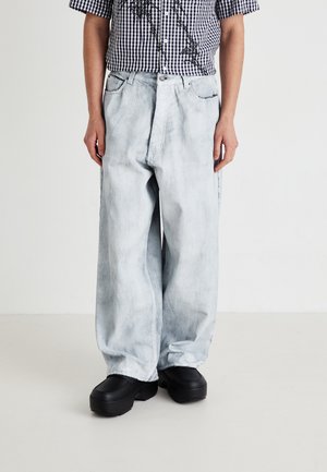 En person som bär ljusvashade vida jeans, svarta plattformsskor och en svart-vitt rutmönstrad kortärmad skjorta med blommönster.