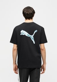 Άτομο που φοράει μαύρο μπλουζάκι με φωτεινό μπλε γραφικό σκελετού τσιτάχ στην πλάτη, στέκεται μπροστά σε απλό ανοιχτό φόντο.