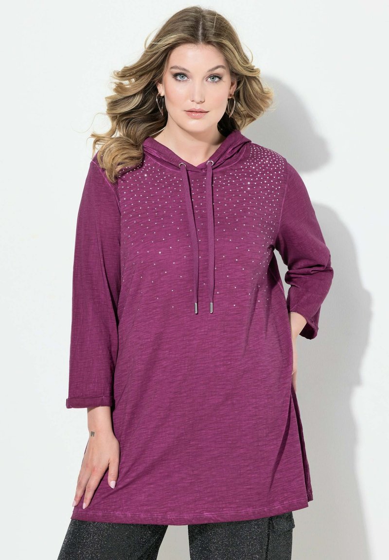Violetti hupullinen tunika, joka on valmistettu teksturoidusta kankaasta. Se on varustettu nyörillä, pitkillä hihoilla ja hopeisten niittien koristeilla edessä.