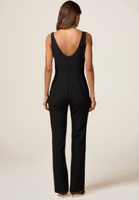Combinaison noire avec un dos en V profond. Le corsage ajusté se prolonge en pantalons évasés, avec une texture lisse et des lignes épurées.