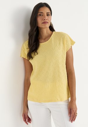 Kvinde med langt mørkt hår læner sig mod en hvid væg, iført en tekstureret gul kortærmet top og hvide bukser, med guldringøreringe.