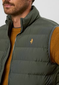 Vest puf verde cu guler înalt și fermoar, având un detaliu cu logo auriu. Purtați peste un pulover texturat maro.