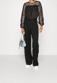 Blusa nera trasparente con decorazioni a fantasia, pantaloni neri a vita alta e una piccola borsa iridescente. Le sneakers bianche completano il look.