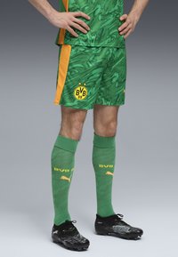 Groene en gele voetbaloutfit met een gestructureerd patroon, zwarte voetbalschoenen en kniehoge groene sokken met gele accenten en logo.