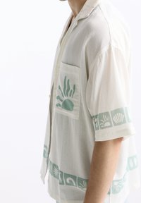 Persona vestida con una camisa corta de color crema con patrones de sol y conchas en turquesa y un bolsillo en el pecho, fotografiada de perfil.