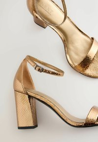 Arany magas sarkú szandálok, texturált kígyóbőr mintával, tömbsarkú, bokapántos kialakítással, fényes felülettel a pántokon és a talpbetéten.