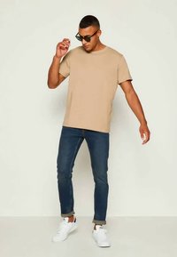 Μπεζ βαμβακερή μπλούζα με στρογγυλή λαιμόκοψη, συνδυασμένη με σκούρα μπλε τζην παντελόνι. Το μοντέλο φορά λευκά αθλητικά παπούτσια και μαύρα γυαλιά ηλίου, στέκεται με άνεση.