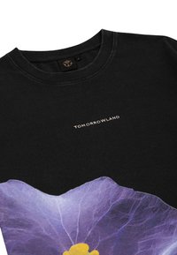 Zwarte katoenen T-shirt met een groot paarse bloemenprint en een gele kern. "TOMORROWLAND" tekst in lichtroze boven het ontwerp.