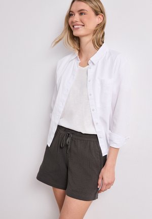 Lächelnde Frau mit blonden Haaren trägt ein weißes Hemd über einem weißen Top und dunkelgraue Shorts mit Kordelzug, stehend vor einem schlichten Hintergrund.