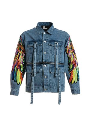 Joven con chaqueta vaquera con alas de lentejuelas de colores en las mangas, camiseta verde tie-dye, pantalones deportivos crema con estampados de alas azules y botas blancas.