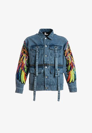 Joven con chaqueta vaquera con alas de lentejuelas de colores en las mangas, camiseta verde tie-dye, pantalones deportivos crema con estampados de alas azules y botas blancas.