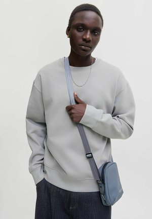 Personne portant un sweat-shirt gris clair, un pantalon foncé, un collier fin en chaîne et un petit sac bandoulière bleu avec une sangle grise sur un fond uni.