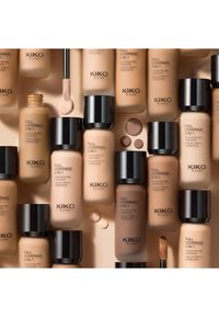 Différentes teintes de fonds de teint et correcteurs 2-en-1 Full Coverage de KIKO Milano avec applicateurs et gouttes de fond de teint sur fond beige.
