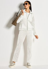 Vrouw in een witte velours trainingspak, met gemusterde sneakers, een grote taupe handtas over haar schouder, oversized zonnebril en gouden oorbellen.