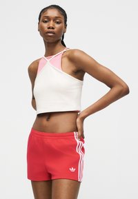 Jeune femme portant un haut court sans manches blanc et rose et un short Adidas rouge avec des bandes blanches, debout avec une main sur la hanche.