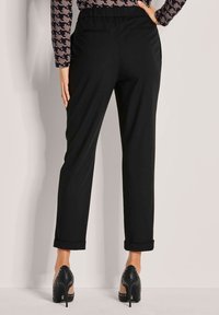 Vrouw in zwarte high-waisted, manchetbroeken en zwarte hoge hakken, staand tegen een lichte muur met één hand op de heup.