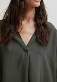 Blusa verde scuro con scollatura a V, maniche corte e un dettaglio a bottone singolo. Realizzata in un tessuto liscio con una vestibilità leggermente ampia.