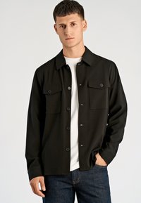 Zwart overhemd met knopen, lange mouwen, twee borstzakken en contrasterende knopen, gecombineerd met een wit T-shirt en donkere jeans.