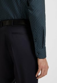 Tummansininen kuvioitu paita, jossa on pieniä vaaleansinisiä geometrisia muotoja; sisältää nappikauluksen ja kontrastoi tummien housujen kanssa, mikä tuo esiin sileän pinnan.