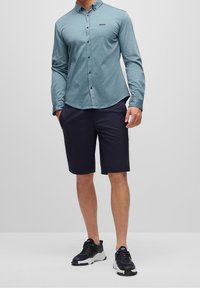 Langärmliges, hellblaues Hemd mit Knöpfen aus leicht strukturiertem Stoff, kombiniert mit dunkelmarineblauen Shorts und schwarzen Sportschuhen.