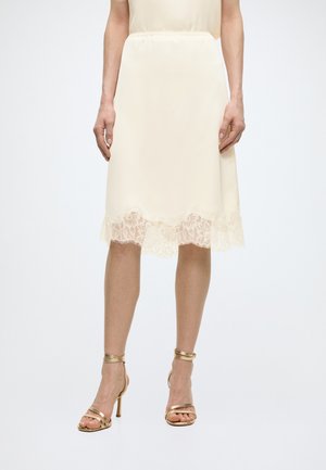 Jupe crème longueur genou avec ourlet en dentelle portée avec des sandales à talons hauts dorées à brides sur jambes nues devant un fond blanc.