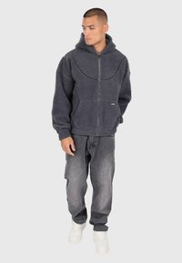 Uomo che indossa un felpa con zip grigia scura, jeans neri sbiaditi e sneakers bianche, in piedi contro uno sfondo semplice.