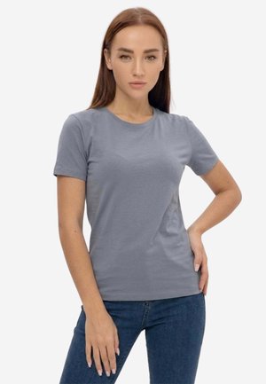 Giovane donna che indossa una semplice t-shirt grigia a maniche corte e jeans blu, in piedi con una mano sul fianco contro uno sfondo bianco.