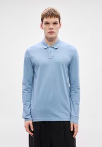 Šviesiai mėlynas ilgomis rankovėmis medvilninis polo marškinėlis su apykakle, dviem mygtukais ir mažu logotipo lopu ant krūtinės.