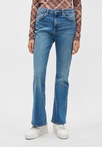 Jeans acampanados de mezclilla en azul claro con cinco bolsillos, cierre de botón y detalles visibles de costura. Combinados con zapatillas blancas.