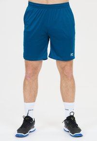 Manliga ben iklädda blå sportshorts, vita crew-strumpor och svarta sportskor som står på vit bakgrund.